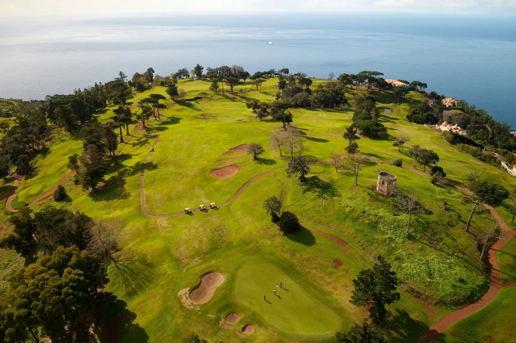 Aerial view of a coastal golf course with greens, bunkers, and scattered trees; ocean appears in the background with buildings along the shore on the right.