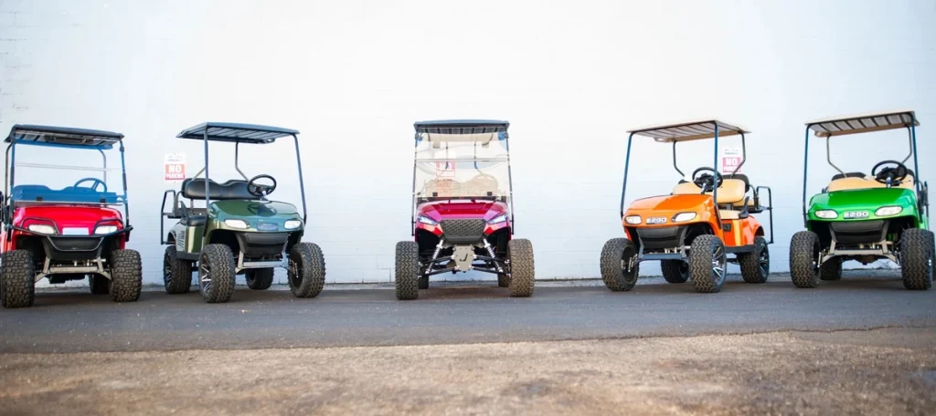 Five lifted golf carts with large tires parked in a row against a plain white wall, each in a different color (red, green, purple, orange, green).