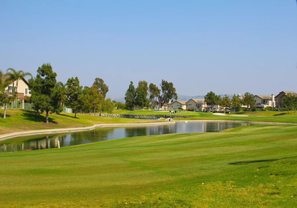 Wide view of a sunny suburban golf course with a pond, green fairways, and houses in the background; a small group walks along the path near the water edge.