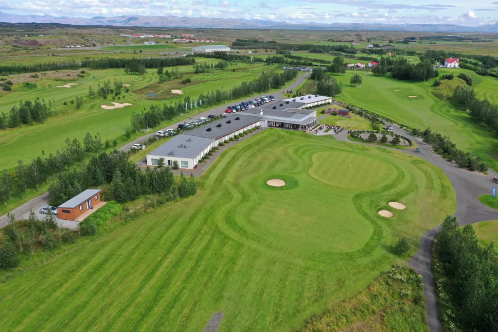Aerial view of a golf course with a long clubhouse building, many parked cars, and green fairways dotted with sand bunkers and trees far and wide.