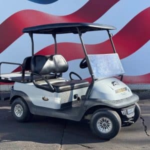 White and gray Club Car golf cart with a black canopy and seats, parked on pavement in front of a red-and-blue wavy mural.