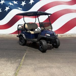 Blue golf cart with tan seats parked on a cracked asphalt surface in front of a large american flag mural.