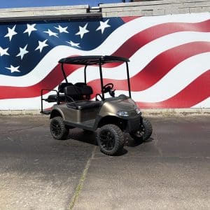 Gray four-seat golf cart with a black roof parked on a paved lot in front of a large red, white, and blue American flag mural.