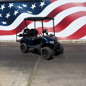 Golf cart with a black roof parked in front of a large American flag mural on a wall, blue body and four seats visible on a sunny day.