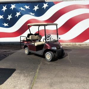 Maroon golf cart with beige seats parked in front of a large american flag mural on a wall.