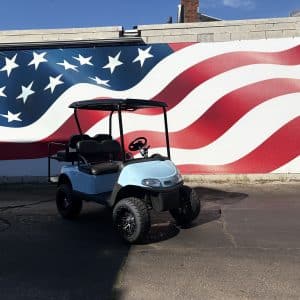 Light-blue golf cart parked in front of a large American flag mural on the wall, with stars and red-and-white stripes.