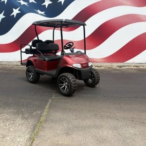 Red golf cart with black seats and a canopy sits on a paved lot in front of a large mural of the american flag.