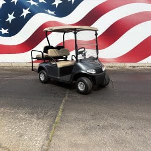 Gray golf cart with tan seats and canopy parked on cracked asphalt in front of a large American flag mural