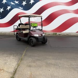 Maroon golf cart with beige seats parked on cracked pavement in front of a large american flag mural, price sign shows $5500.