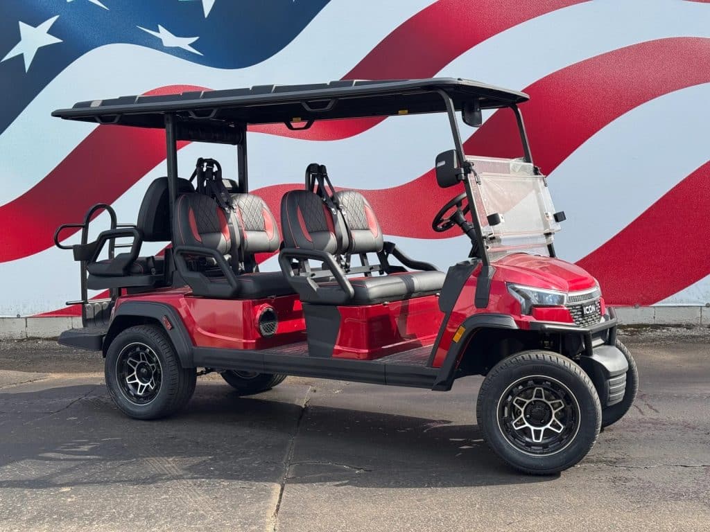 Six-passenger red golf cart with black and red diamond-stitched seats parked in front of a large American flag mural, under a black roof.