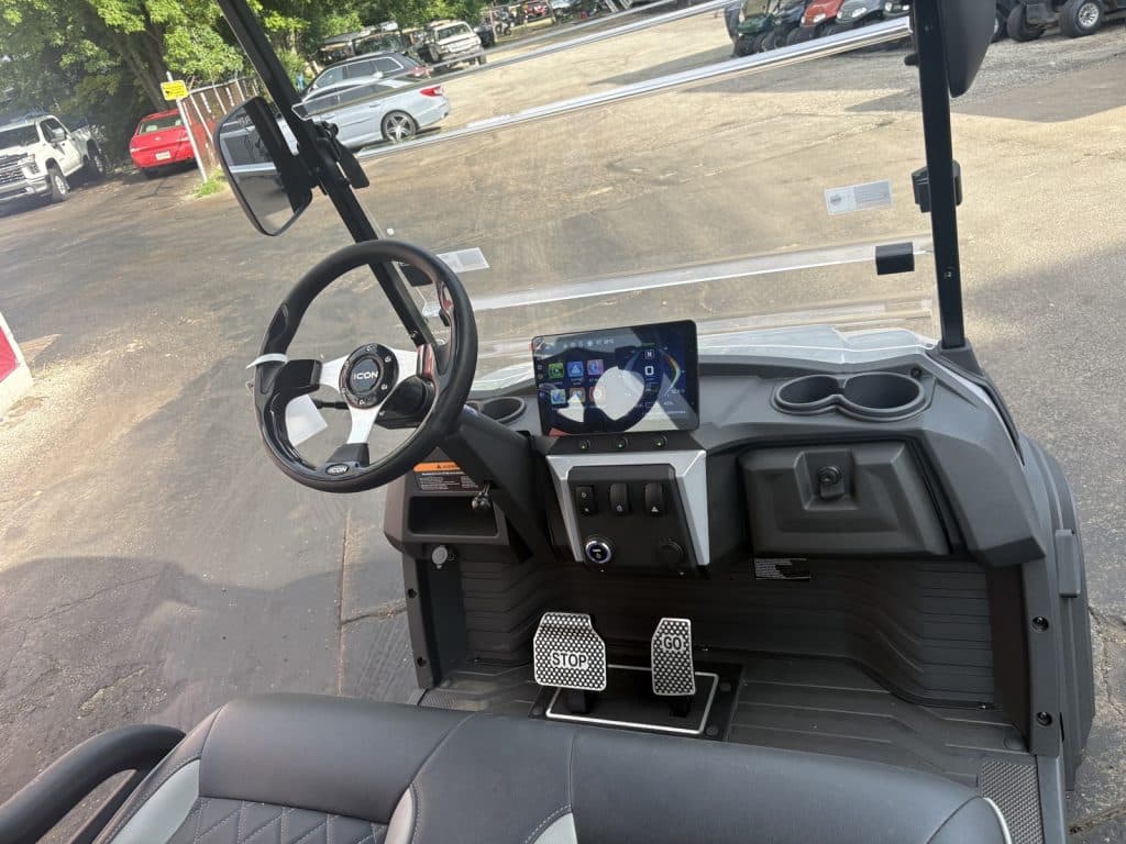 Cockpit of a golf cart with a touchscreen infotainment panel, icon steering wheel, two cup holders, and labeled stop/go pedals in front of the seat.