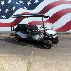 Light blue golf cart parked on a pavement in front of a large mural of the american flag with white stars on blue and red stripes. Four black leather seats visible under a canopy.