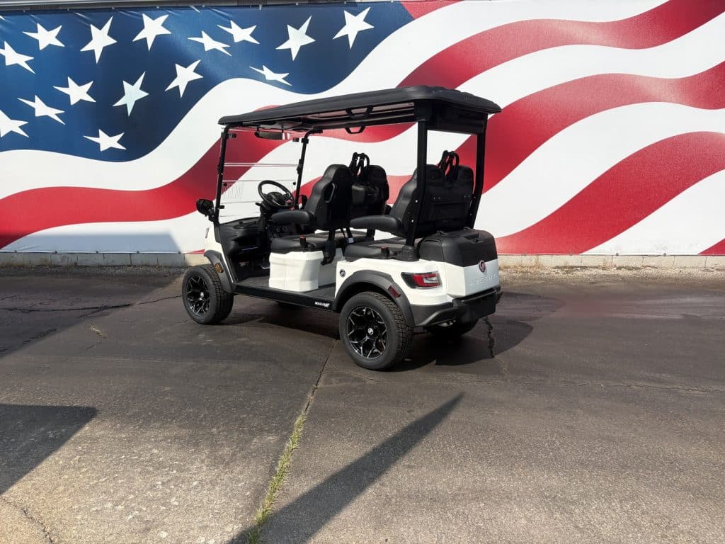 White golf cart with a black roof parked on a paved lot in front of a large American flag mural with red, white, and blue waves and stars.