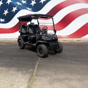 Black electric golf cart with four seats parked on a cracked pavement in front of a large wall mural of the american flag with red and white stripes and white stars on blue.