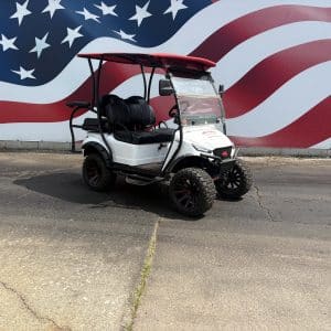 White golf cart with a red roof and rugged tires in front of a large american flag mural.