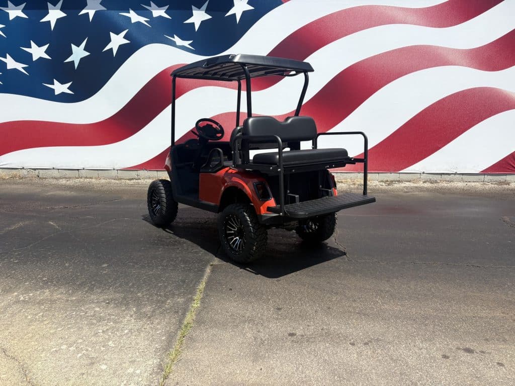Red lifted golf cart with black seats and a rear cargo platform parked on a parking lot in front of a large American flag mural behind it.