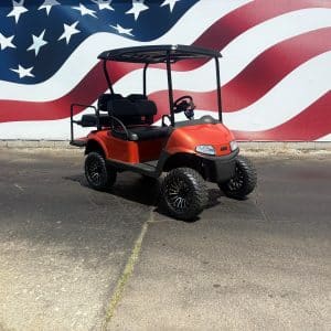 Red golf cart with large off-road tires parked on a pavement in front of a large American flag mural.