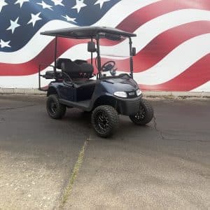 Blue golf cart with a black roof parked in front of a large american flag mural in a paved lot.