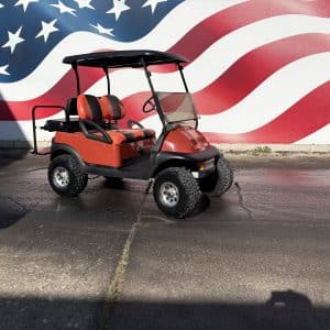 Red golf cart parked in front of a large American flag mural, with orange-and-black seats and a black roof.