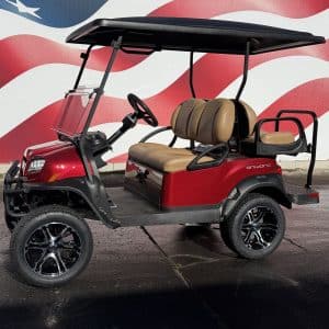 Red golf cart with tan seats and black roof parked on a pavement with a large American flag mural in the background; windshield visible.